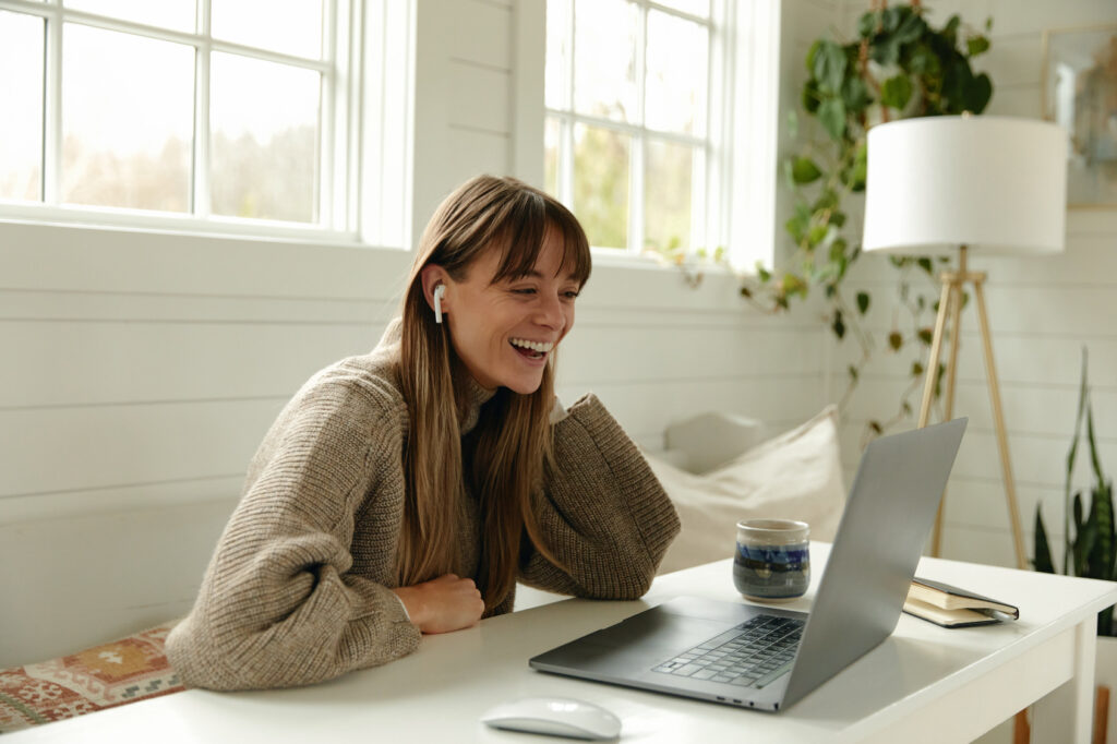A woman with headphones on is at a desk smiling and communicating with someone online while a coffee sits on the desk.